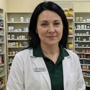 Slightly smiling woman with black hair, wearing a lab coat and standing in a pharmacy